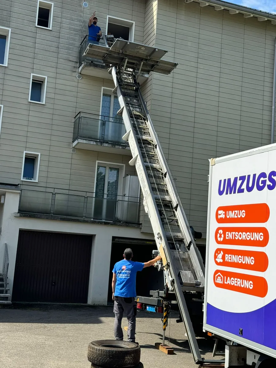 Möbellift an der Hausfassade: ein Mitarbeiter bedient den Lift, ein zweiter nimmt auf dem Balkon entgegen